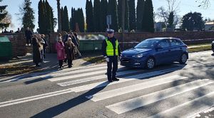 Policjant stoi na przejściu dla pieszych i kieruje ruchem. Z boku stoi auto, przed przejściem piesi.