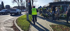 Policjant idzie chodnikiem przy cmentarzu. Wokół idą ludzie, na drodze jadą auta.