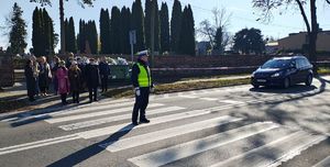 Policjant stoi na przejściu dla pieszych i kieruje ruchem. Z boku stoi auto, przed przejściem piesi.