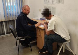 Zatrzymany mężczyzna siedzi przy biurku i podpisuje dokumenty, obok siedzi policjant.