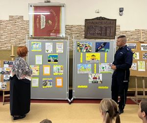 Hol w Szkole Podstawowej numer 4 w Wieluniu, na sztalugach prezentowane są prace plastyczne konkursowe, z boku stoją pani dyrektor oraz policjant.