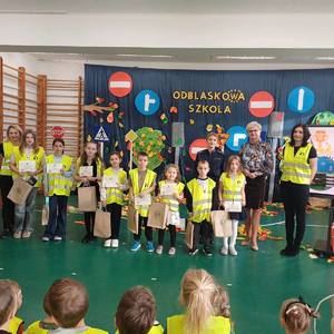 Sala gimnastyczna w Szkole Podstawowej w Bieniadzicach, dyrekcja, nauczyciele, policjantka wspólnie z laureatami konkursu. Dzieci ubrane są w kamizelki odblaskowe.