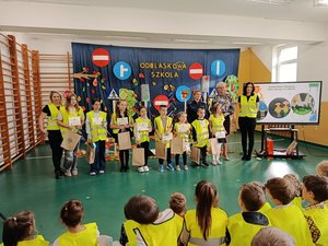 Sala gimnastyczna w Szkole Podstawowej w Bieniadzicach, dyrekcja, nauczyciele, policjantka wspólnie z laureatami konkursu. Dzieci ubrane są w kamizelki odblaskowe.