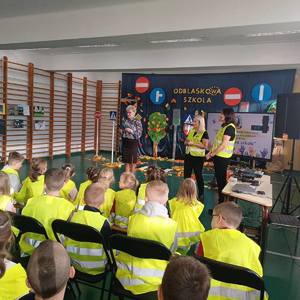 Sala gimnastyczna w Szkole Podstawowej w Bieniadzicach, na krzesłach siedzą dzieci ubrane w kamizelki odblaskowe, na środku przed dziećmi stoi pani dyrektor oraz nauczycielki.