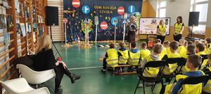 Sala gimnastyczna w Szkole Podstawowej w Bieniadzicach, na krzesłach siedzą dzieci ubrane w kamizelki odblaskowe, na środku przed dziećmi stoi pani dyrektor oraz nauczycielki.