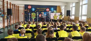 Sala gimnastyczna w Szkole Podstawowej w Bieniadzicach, na krzesłach siedzą dzieci ubrane w kamizelki odblaskowe, na środku przed dziećmi stoi pani dyrektor oraz nauczycielki.