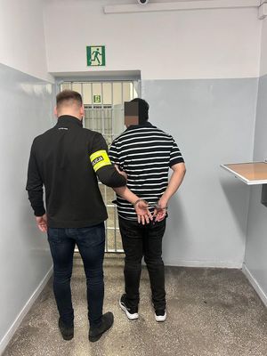 Policjant prowadzi zatrzymanego po korytarzu w budynku komendy.