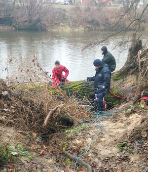 Strażak wychodzi z wody, na brzegu rzeki znajduje się drugi strażak i policjant.
