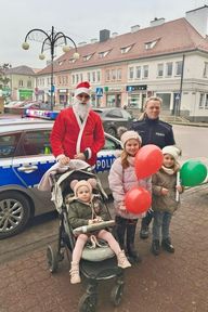 Radiowóz, przed nim stoi policjantka, osoba w stroju Mikołaja oraz dzieci, jedno jest w wózku.Dzieci trzymają w ręku balony.