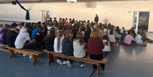 Policjantka prowadzi prelekcję w sali gimnastycznej gdzie dzieci siedzą na ławkach.