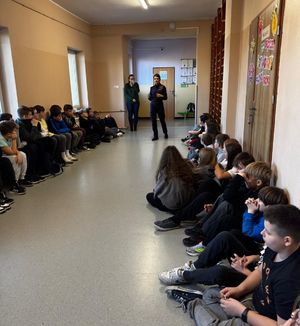 Policjantka prowadzi prelekcję na korytarzu w szkole, z boku siedzą dzieci.