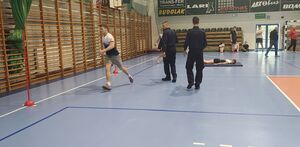 Hala sportowa na której rozstawiony jest tor przeszkód, zawodnicy wykonują ćwiczenia sportowe. Policjanci nadzorują zawody.