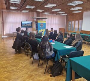 Sala odpraw w wieluńskiej jednostce Policji, przy stole siedzą uczniowie i policjant, na ekranie wyświetlony film.