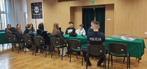 Sala odpraw w wieluńskiej jednostce Policji, przy stole siedzą uczniowie i policjant prowadzący rozmowę.
