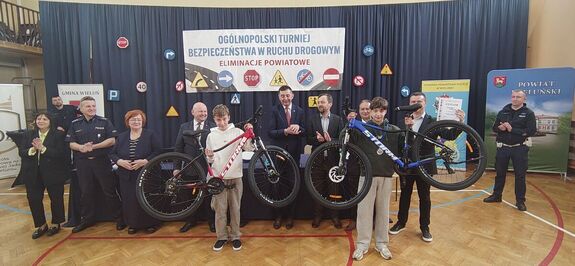 Sala gimnastyczna w Szkole Podstawowej numer 4 w Wieluniu. Zaproszeni goście, funkcjonariusze  oraz dwóch laureatów konkursu prezentuje dwa rowery, które stanowiły nagrody.