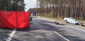 Droga z boku stoi w rowie uszkodzony samochód, po drugiej stronie parawan straży pożarnej. W oddali na drodze stoi wóz strażacki.