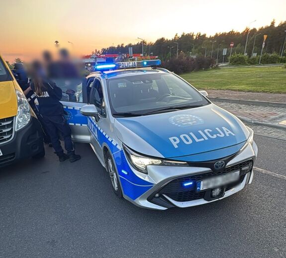 Radiowóz policyjny na drodze. policjantka stoi przy radiowozie z innymi uczestnikami interwencji, z boku widać element skradzionego pojazdu.