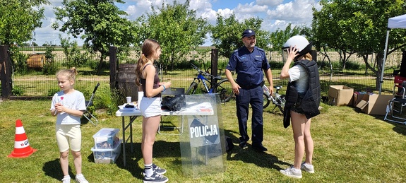 Policjant na stoisku profilaktycznym przy którym stoją dzieci. Jedno z dzieci przymierza elementy umundurowania policjanta, chełm, kamizelkę.