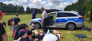 Uczestnicy turnieju oglądają elementy wyposażenia policyjnego, radiowóz, w tle stoi policjant.