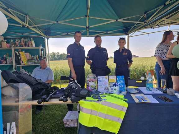 Policjanci na pikniku pod namiotem na wystawionym stosiku profilaktycznym prezentują sprzęt policyjny, na stole wyłożone elementy odblaskowe, ulotki i drobne gadżety promocyjne.