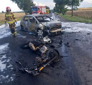 Droga na której stoi spalony samochód i motocykl, obok stoi strażak.