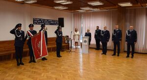 Sala odpraw wieluńskiej jednostki Policji, w sali stoją Zastępca Komendanta Wojewódzkiego Policji w Łodzi, Komendant Powiatowy Policji w Wieluniu, I Zastępca Komendanta Powiatowego Policji w Wieluniu.  Z boku poczet sztandarowy.