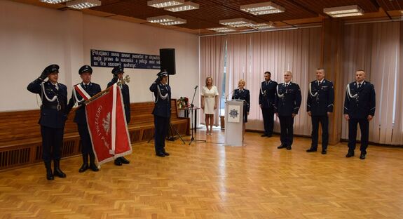 Sala odpraw wieluńskiej jednostki Policji, w sali stoją Zastępca Komendanta Wojewódzkiego Policji w Łodzi, Komendant Powiatowy Policji w Wieluniu, I Zastępca Komendanta Powiatowego Policji w Wieluniu.  Z boku poczet sztandarowy.