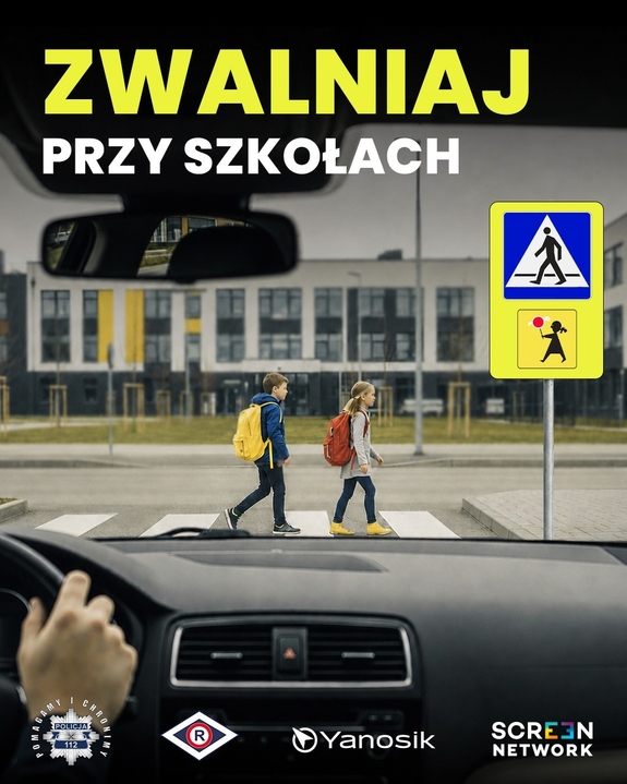 Wnętrze a, kierownica i ręce trzymane na niej. Z wnętrza pojazdu widać przejście dla pieszych, idące po nim dzieci i znaki drogowe informujące o przejściach dla pieszych. Napis zwalniaj przy szkołach.