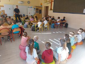 Sala lekcyjna policjant prowadzi pogadankę z dziećmi, które siedzą w ławkach.