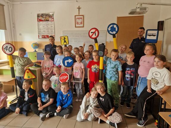 Sala lekcyjna, policjanci stoją w rzędach z dziećmi, które prezentują znaki drogowe.