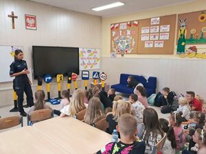 Sala lekcyjna policjantka prowadzi pogadankę z dziećmi, które siedzą w ławkach.