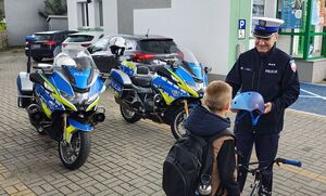 Policjant z ruchu drogowego stoi na parkingu, trzyma w ręku  kask ochrony, na przeciwko nich stoi chłopiec z rowerem. Na parkingu stoją zaparkowane motocykle policyjne.