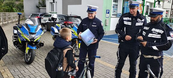 Policjanci z ruchu drogowego stoją na chodniku , jeden z policjantów trzyma w ręku pudełko, w którym jest kask ochronny, na przeciwko nich stoi chłopiec z rowerem.