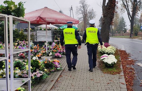 Rejon cmentarza, z boku stoją kwiaty, policjanci idą chodnikiem.